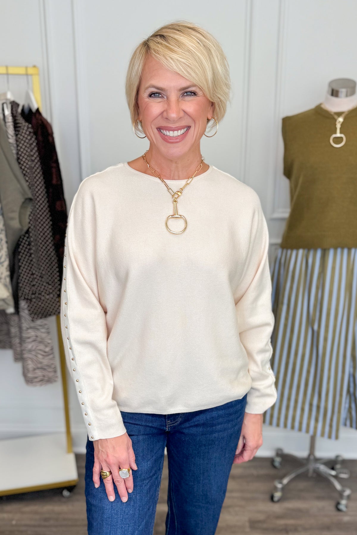 Front view of woman wearing Swagger Boutique’s Ecru Studded Sleeve Sweater  with jeans with clothing hanging in the background.