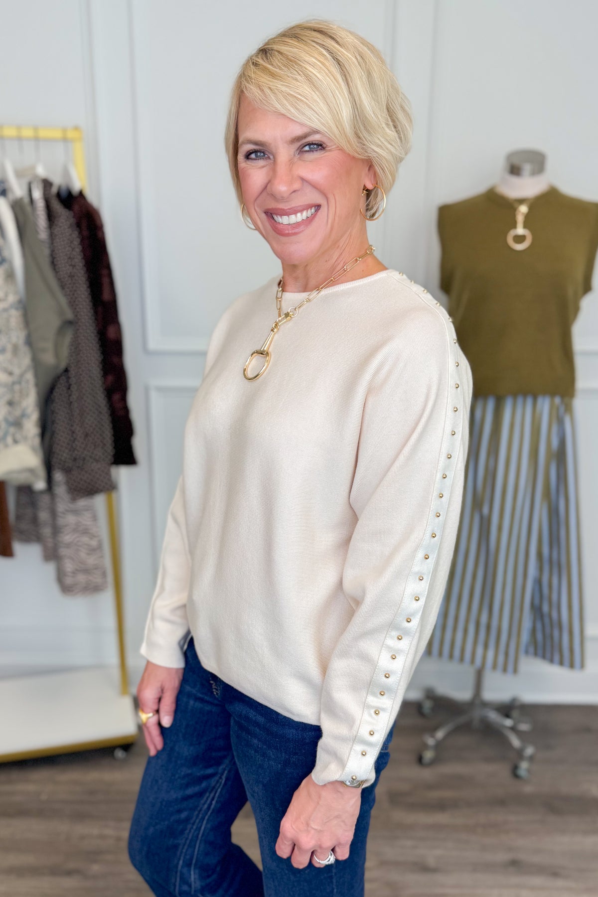 Side view of woman wearing Swagger Boutique’s Ecru Studded Sleeve Sweater  with jeans with clothing hanging in the background.