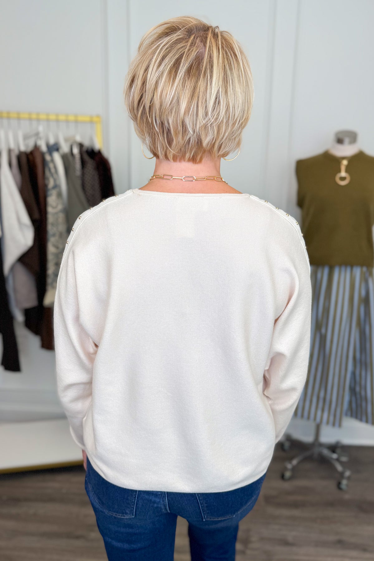 Back view of woman wearing Swagger Boutique’s Ecru Studded Sleeve Sweater  with jeans with clothing hanging in the background.