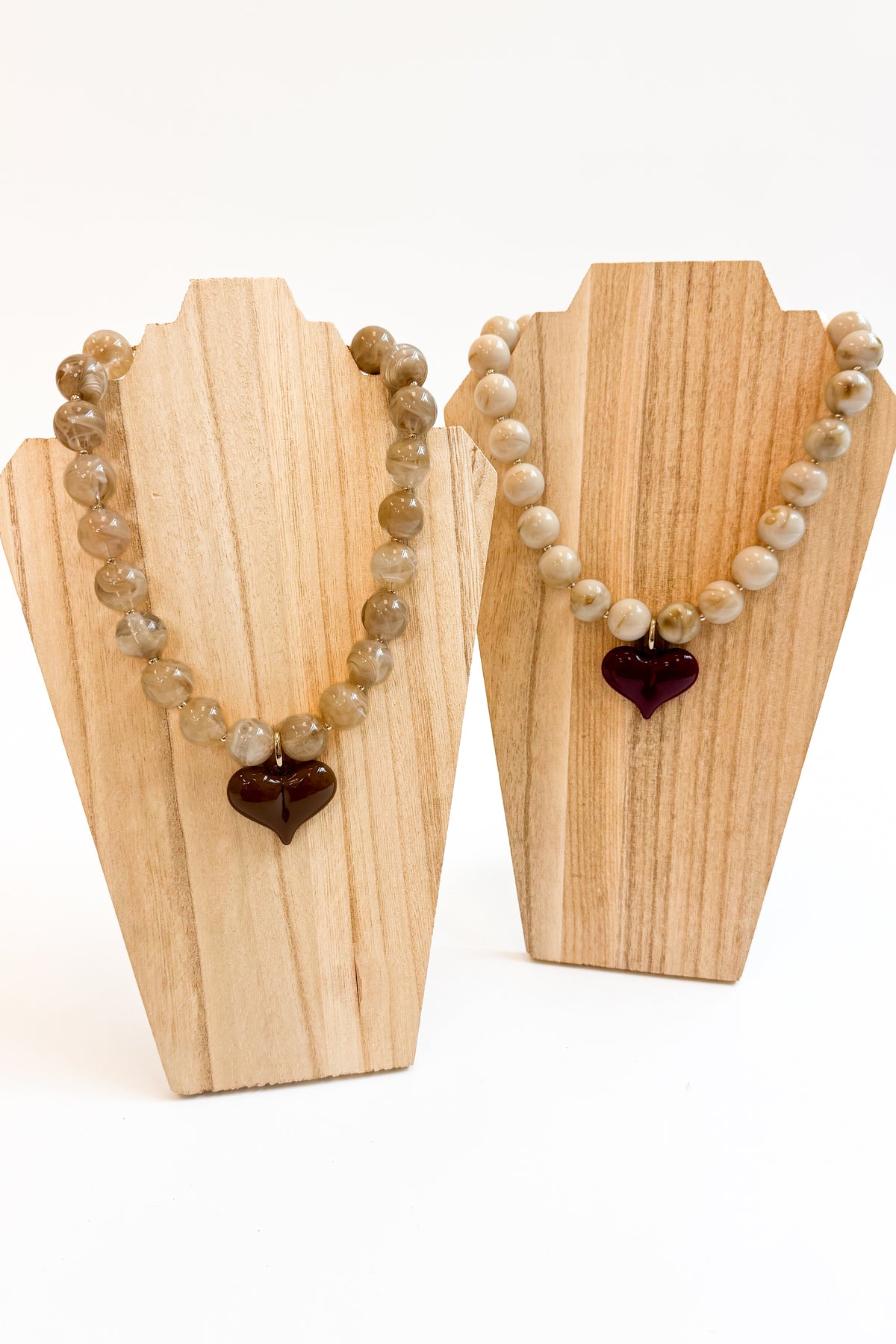 Two beaded necklaces displayed on light wood standees against a white background. One necklace features round tan and gray marbled beads with a dark brown heart-shaped pendant.