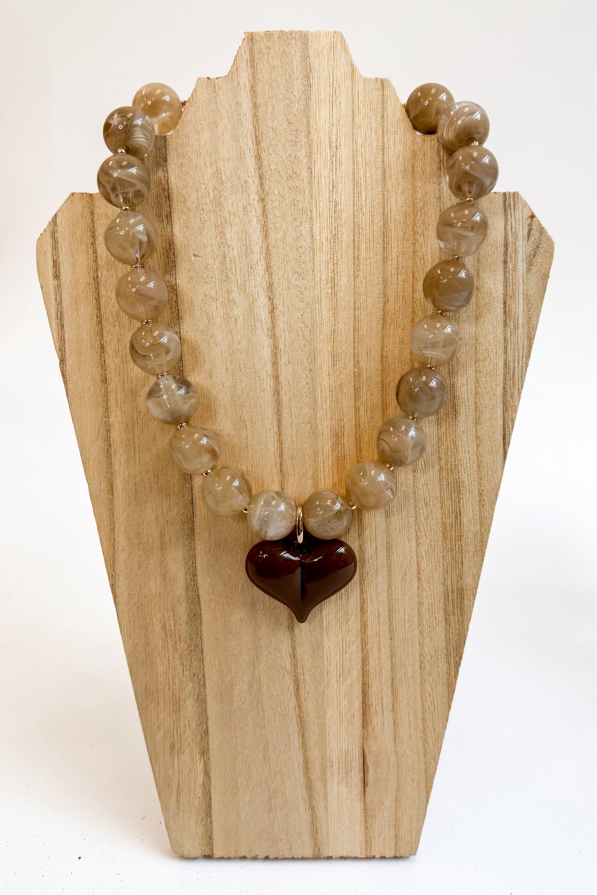 A beaded necklace made of round, translucent light brown and beige gemstone-like beads with a dark reddish-brown heart-shaped pendant, displayed on a light wood stand against a white background.