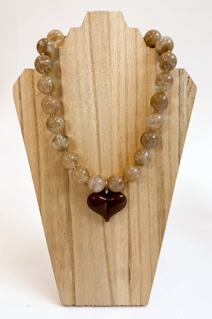 A beaded necklace made of round, translucent light brown and beige gemstone-like beads with a dark reddish-brown heart-shaped pendant, displayed on a light wood stand against a white background.