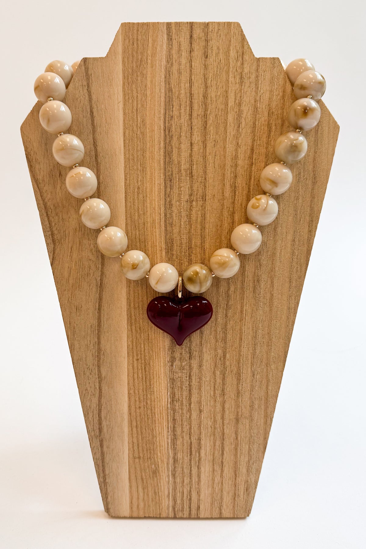 A beaded necklace made of cream and tan marbled beads with a glossy dark red heart-shaped pendant, displayed on a wooden stand against a white background.