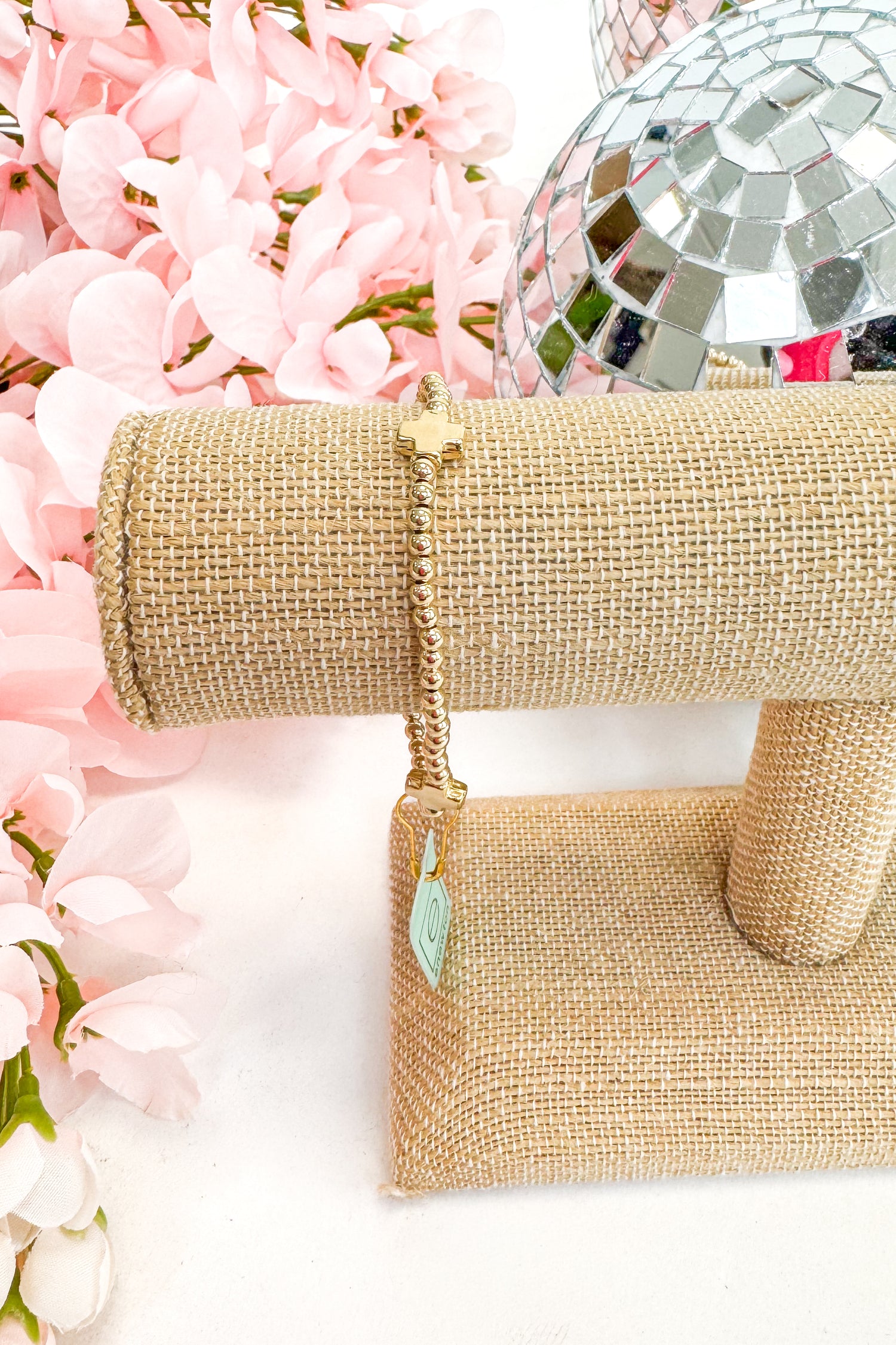 gold bracelet with gold cross detail on a white background with pink flowers and a disco ball.