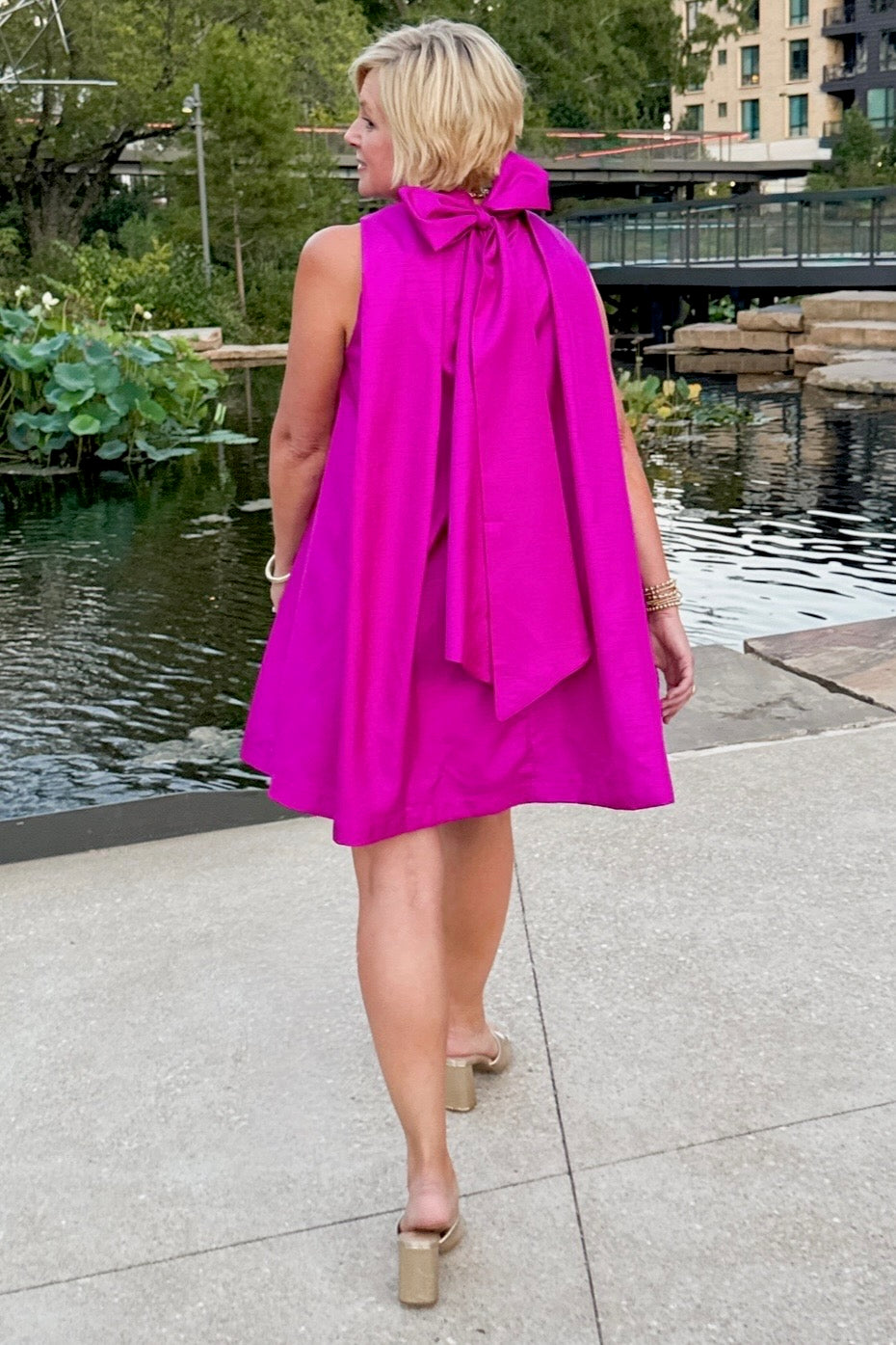 Woman wearing a bright pink dress with a bow detail, walking by a water body.