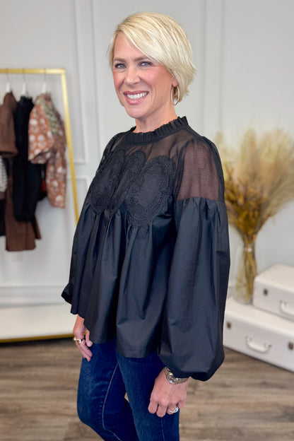 Woman wearing a black blouse with sheer sleeves in a room with clothing and decor.