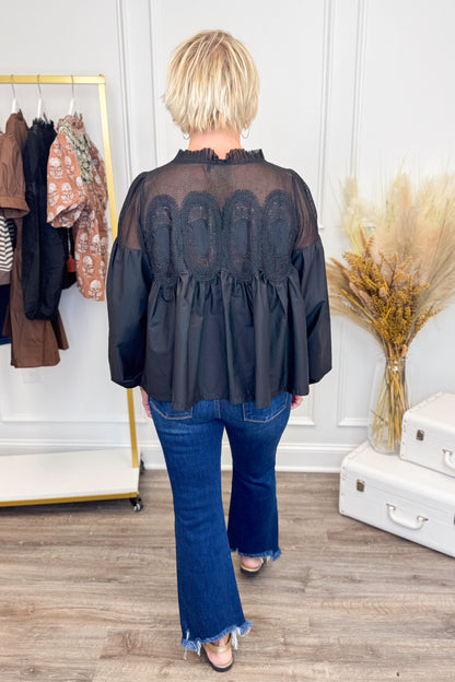 Person wearing a black blouse with a pattern and blue jeans in a room with a coat rack and decorative items.