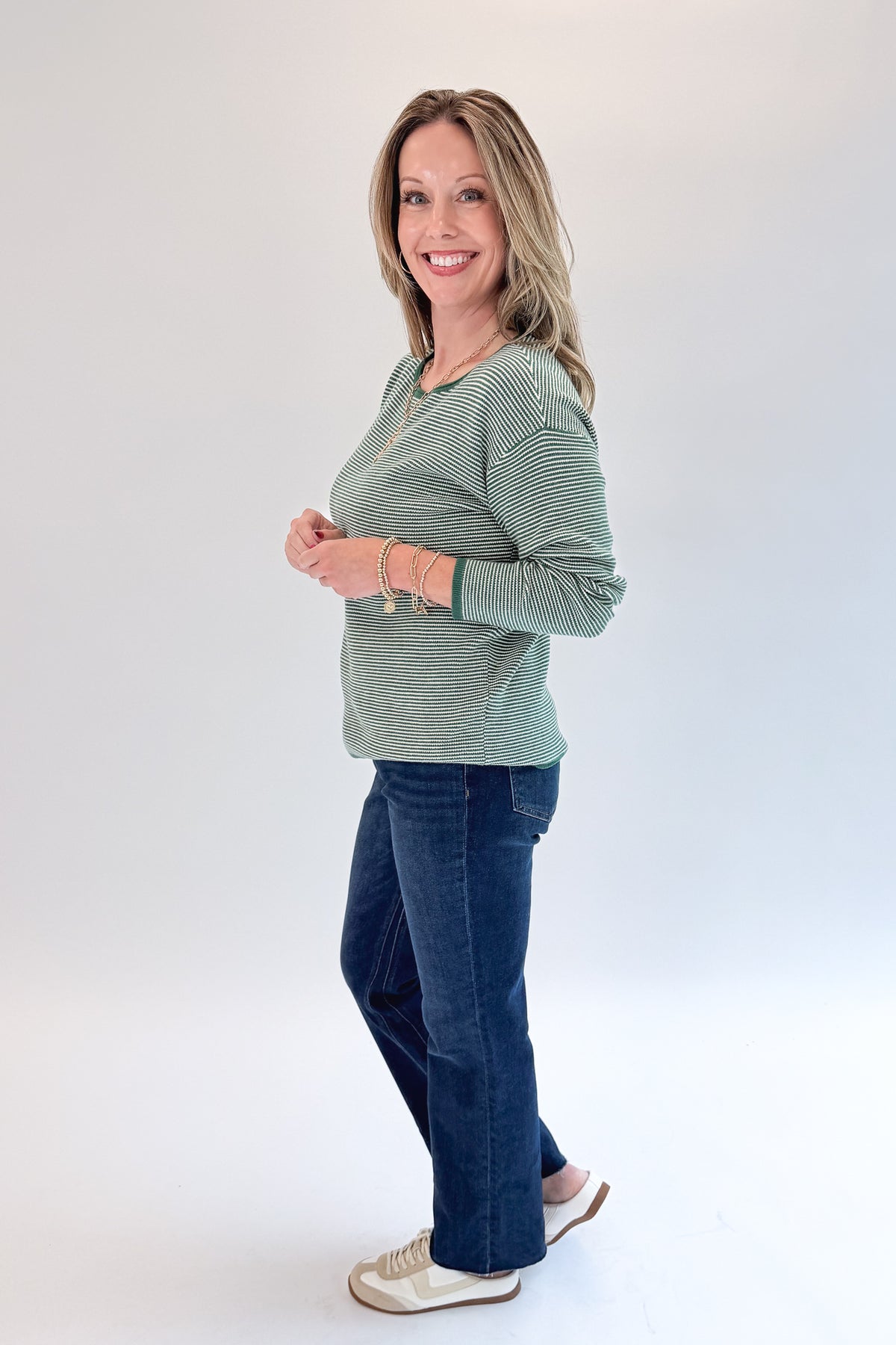 Side view of woman wearing Swagger Boutique’s Forest Green Striped Sweater with jeans against white studio background.