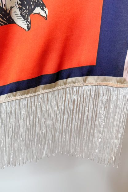A close-up of a scarf featuring a bright orange field, a navy blue border, a white fringe trim, and part of a black-and-white horse graphic near the top left.