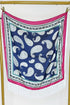 A square scarf with a navy blue background, featuring a white paisley pattern, a pink border, and white fringe at the bottom, hanging from a gold-colored metal stand against a white wall.