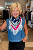 A smiling blonde woman wearing a blue denim sleeveless top and a colorful fringed neck scarf with white fringe, standing inside a clothing boutique.