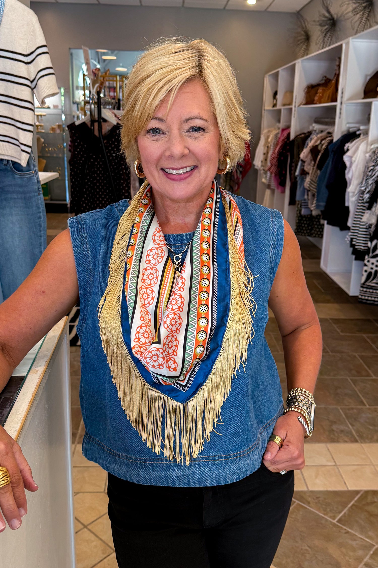 A smiling blonde woman wearing a blue denim vest over a colorful patterned scarf with long cream fringe, standing inside a clothing store.