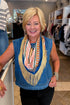A smiling blonde woman wearing a blue denim vest over a colorful patterned scarf with long cream fringe, standing inside a clothing store.