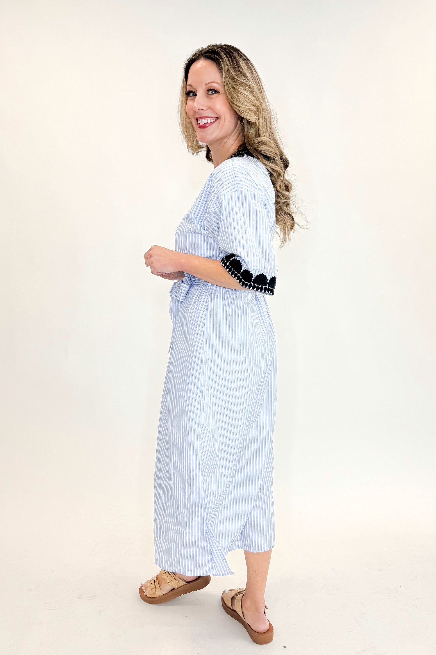 Side view of woman wearing Blue Striped Embroidery Accent Midi Dress with sandals against white studio background.