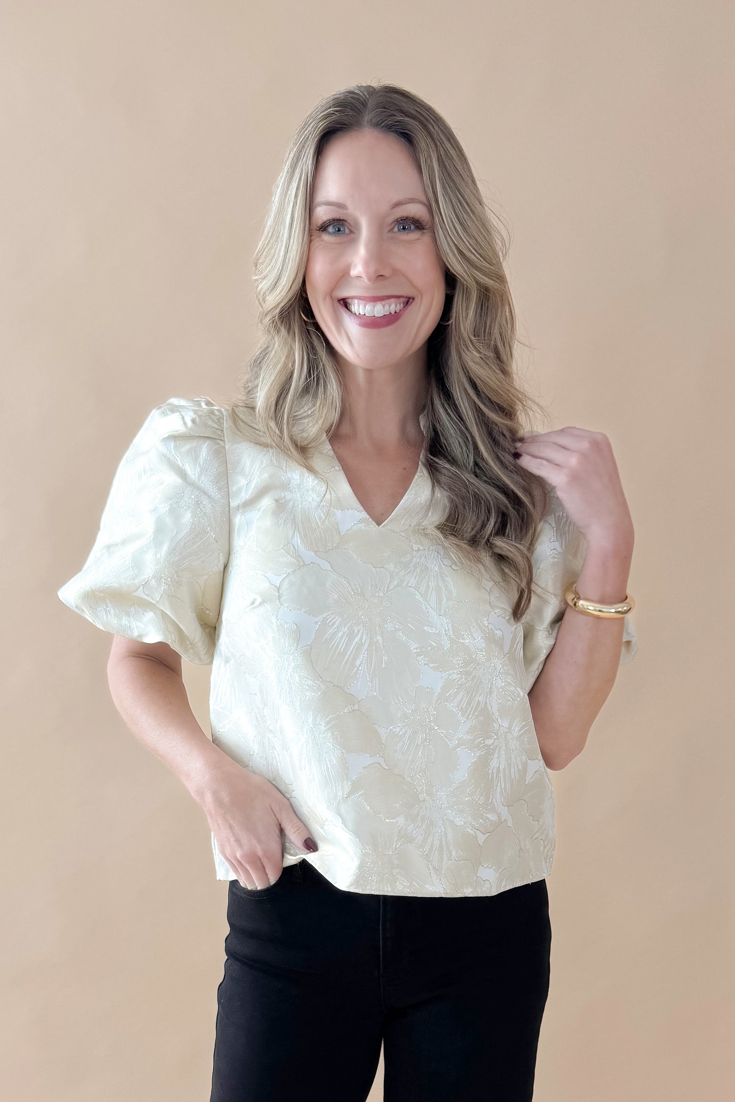 Front view of woman wearing Champagne Metallic Textured Floral Top with jeans against tan studio background.