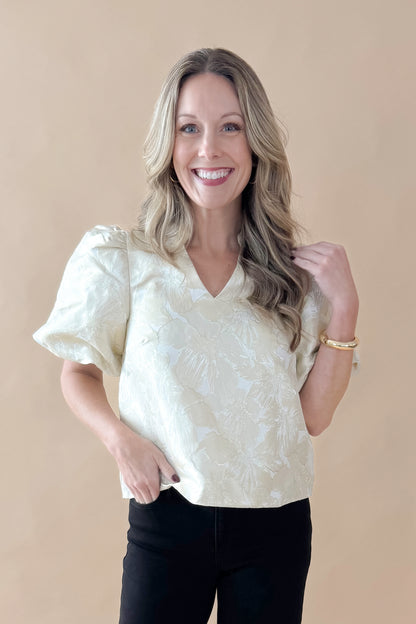Front view of woman wearing Champagne Metallic Textured Floral Top with jeans against tan studio background.