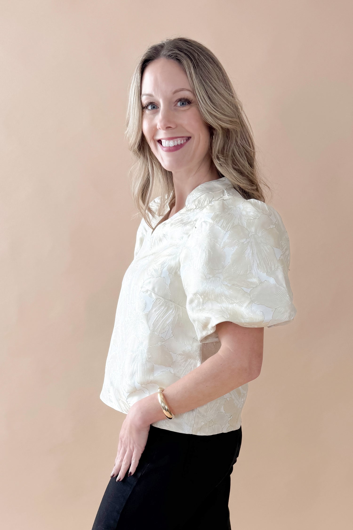 Side view of woman wearing Champagne Metallic Textured Floral Top with jeans against tan studio background.