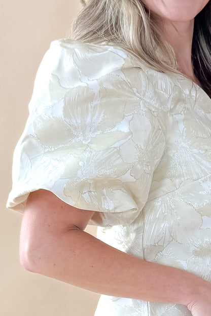 Side detailed view of woman wearing Champagne Metallic Textured Floral Top with jeans against tan studio background.