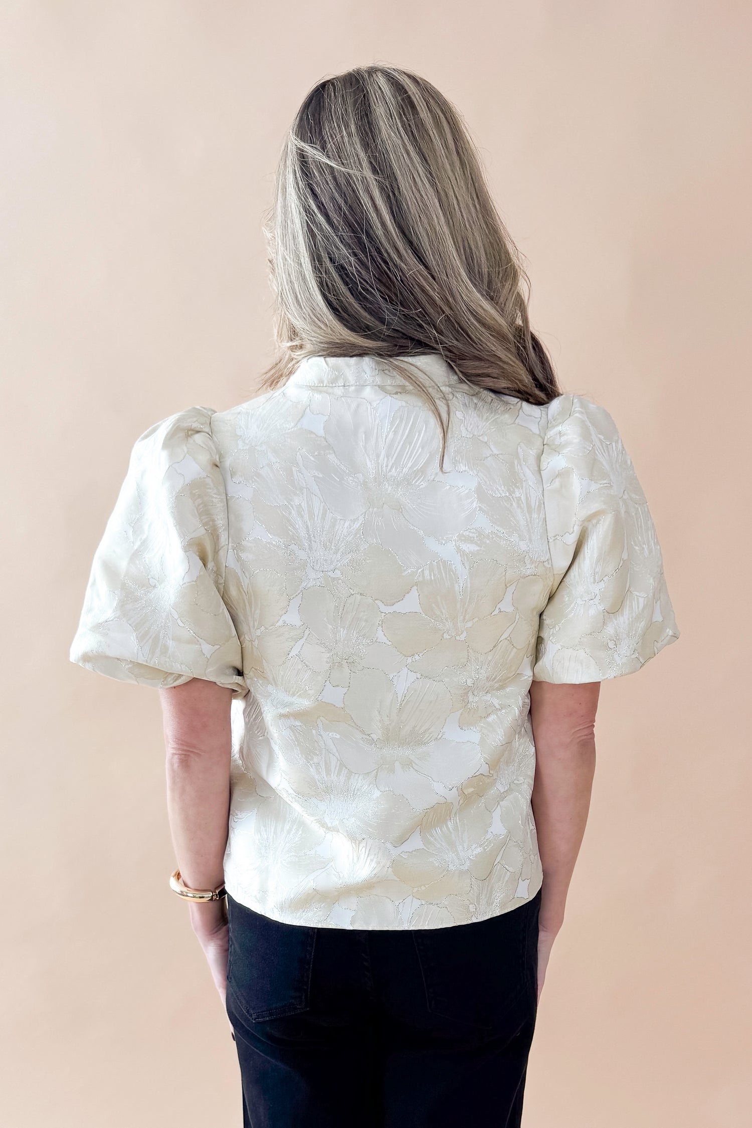 Back view of woman wearing Champagne Metallic Textured Floral Top with jeans against tan studio background.