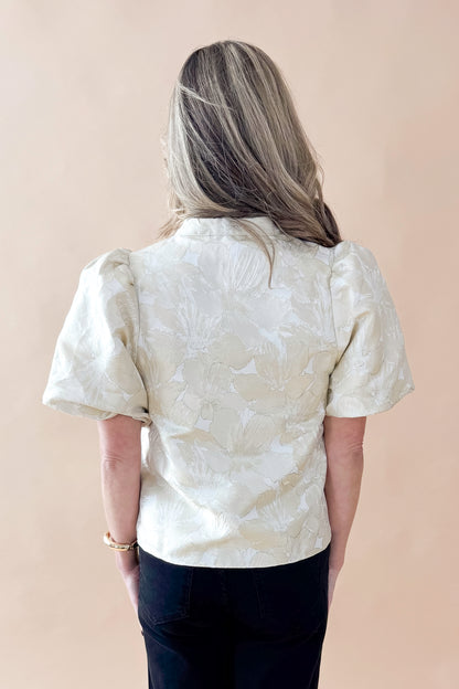 Back view of woman wearing Champagne Metallic Textured Floral Top with jeans against tan studio background.