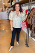 Front view of woman wearing Champagne Metallic Textured Floral Top with jeans at boutique with clothes in background.