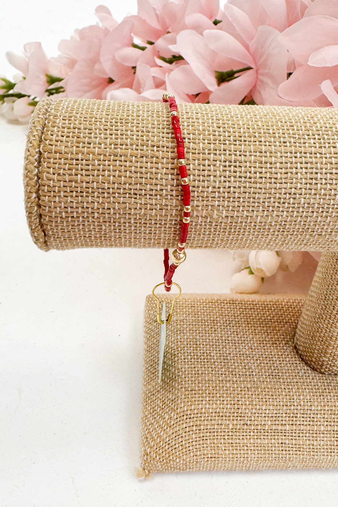 Red beaded bracelet with a gold tag on a beige mesh stand with pink flowers in the background