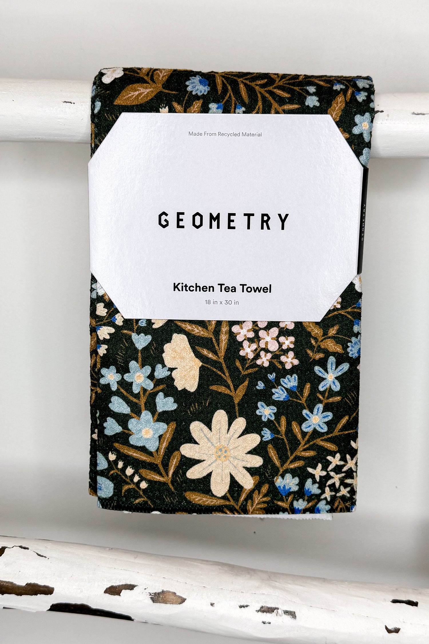 A kitchen tea towel with a black background and a botanical floral pattern in cream, light blue, and gold, hanging from a white painted wooden rod. A white paper label is attached to the center with the text &