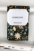 A kitchen tea towel with a black background and a botanical floral pattern in cream, light blue, and gold, hanging from a white painted wooden rod. A white paper label is attached to the center with the text &