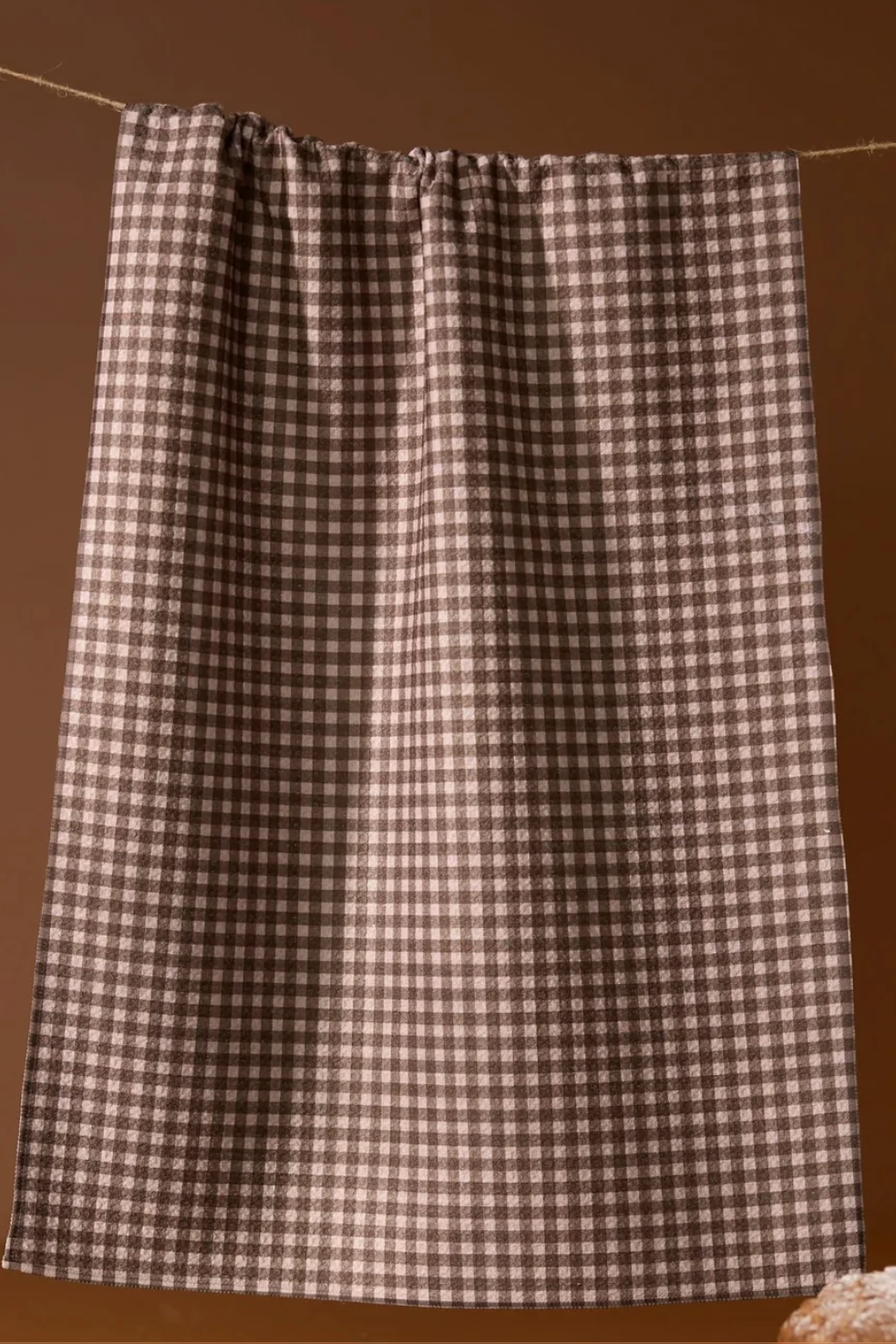 A brown and beige gingham towel  hanging from a rope against a solid brown background.