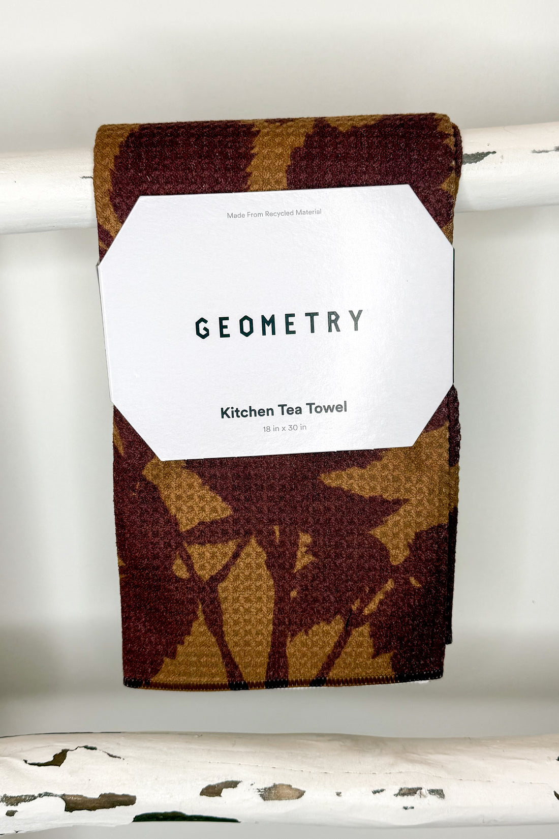 A folded kitchen tea towel with a brown and tan waffle-weave pattern hangs from a white rod. A white paper label on the front reads &