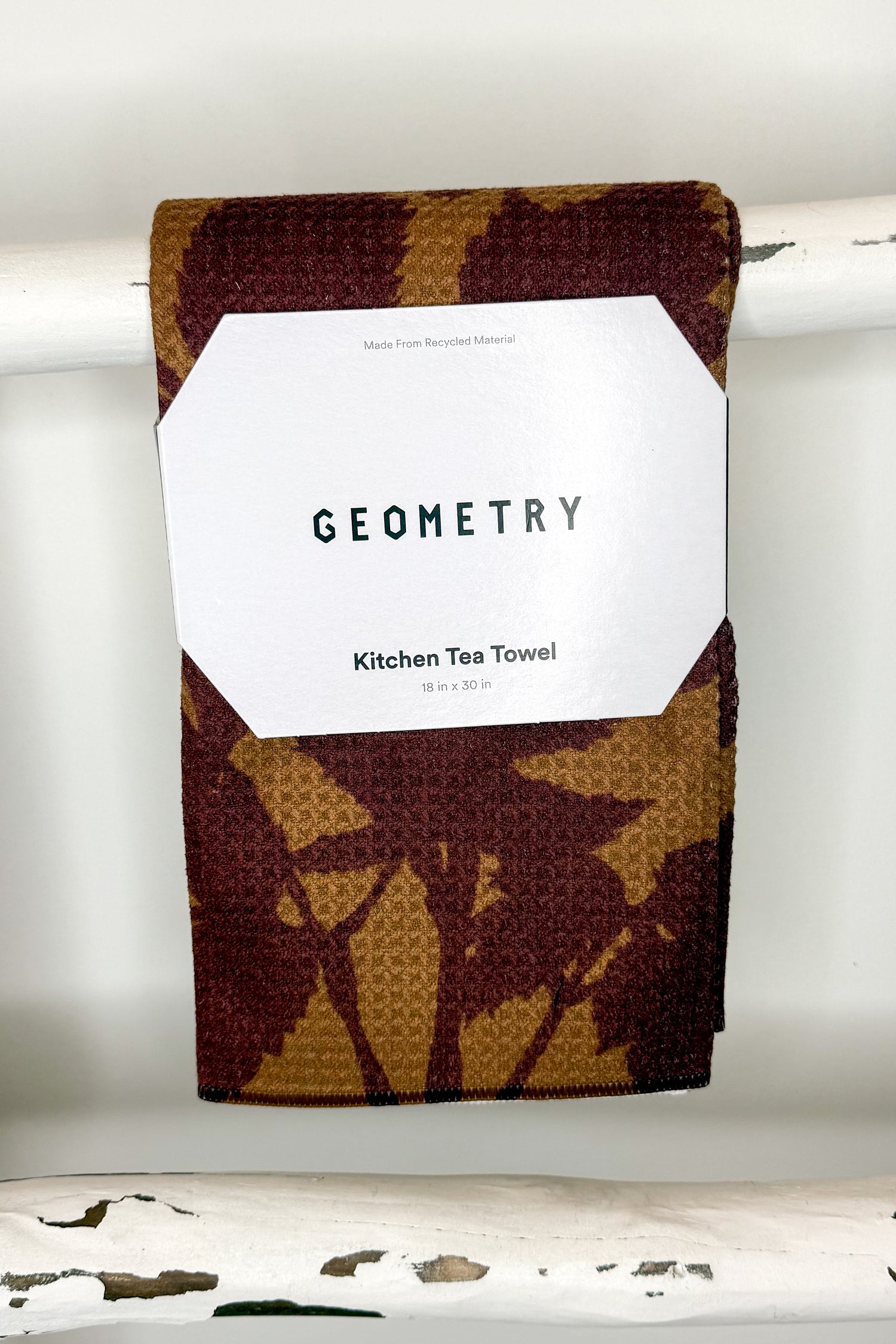 A folded kitchen tea towel with a brown and tan waffle-weave pattern hangs from a white rod. A white paper label on the front reads &