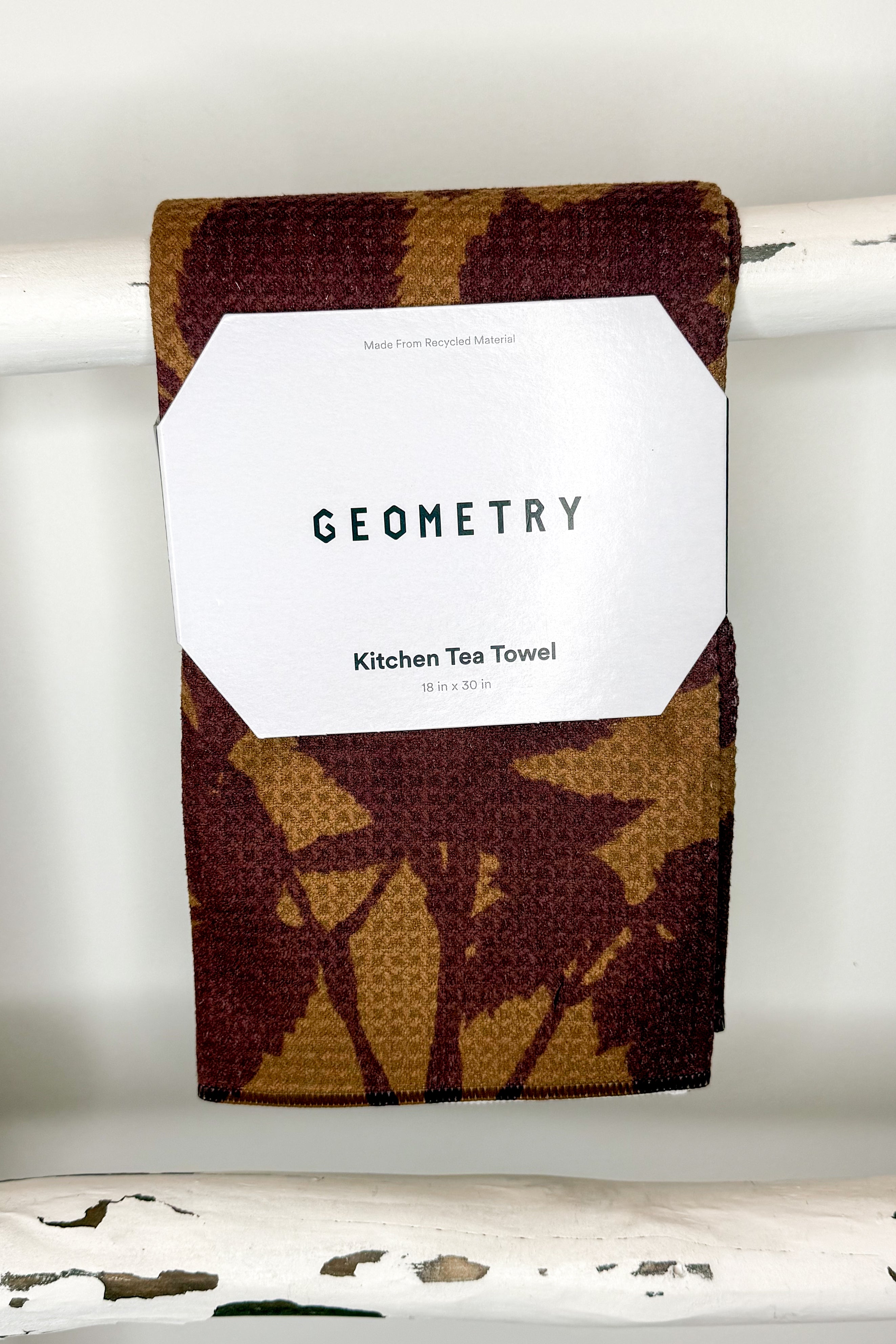 A folded kitchen tea towel with a brown and tan waffle-weave pattern hangs from a white rod. A white paper label on the front reads &
