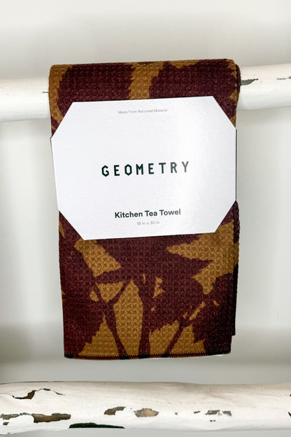 A folded kitchen tea towel with a brown and tan waffle-weave pattern hangs from a white rod. A white paper label on the front reads &