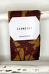 A folded kitchen tea towel with a brown and tan waffle-weave pattern hangs from a white rod. A white paper label on the front reads &