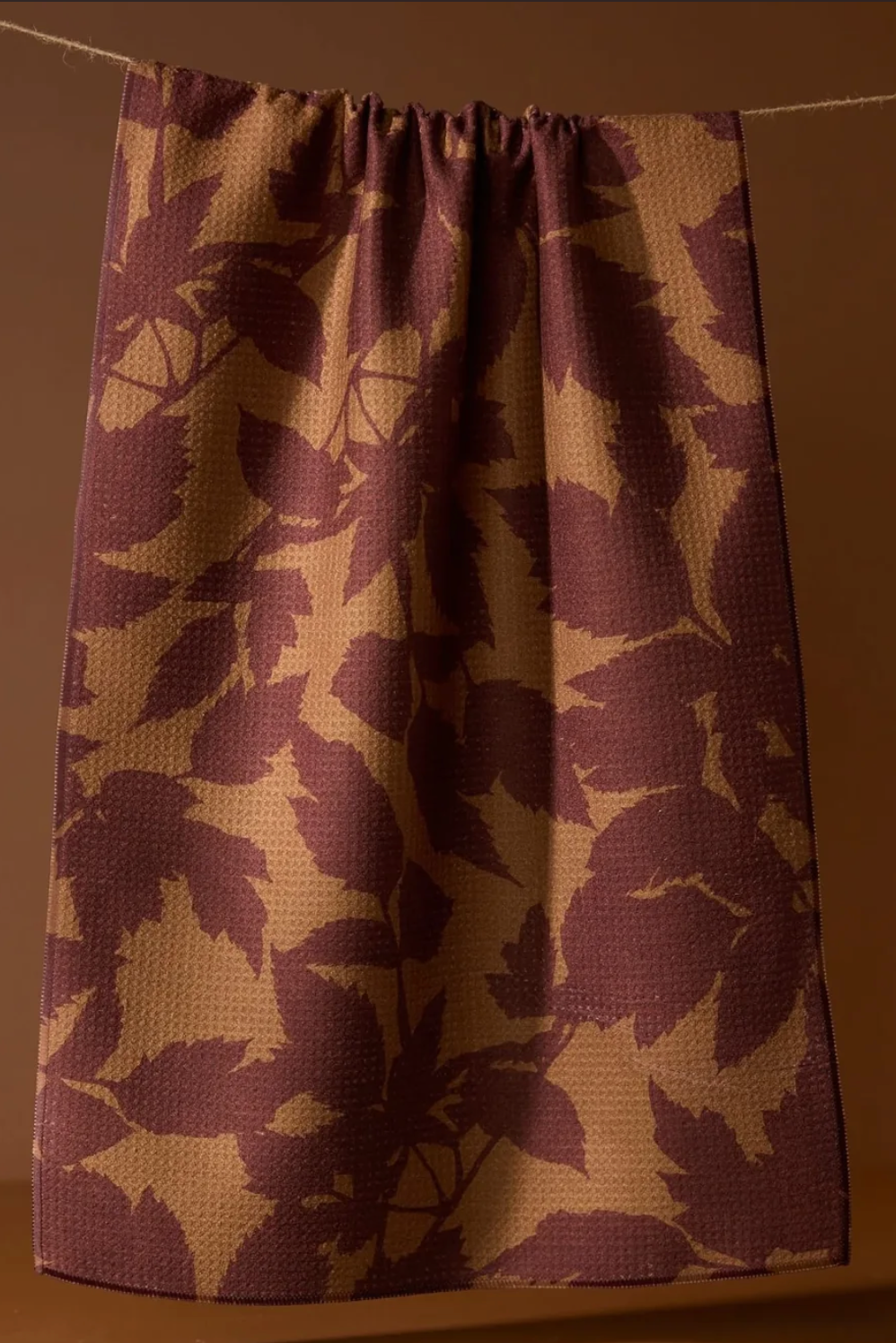 A towel with a waffle-weave texture, featuring a dark plum purple and tan leaf pattern, hanging from a thin rope against a solid warm brown background.