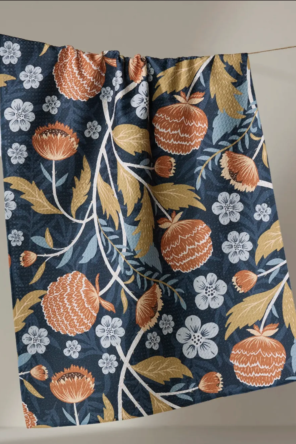 A hanging towel with a botanical print featuring orange pomegranate-like fruits, gold leaves, white and blue-gray flowers, and a dark navy background, suspended against a plain beige wall.