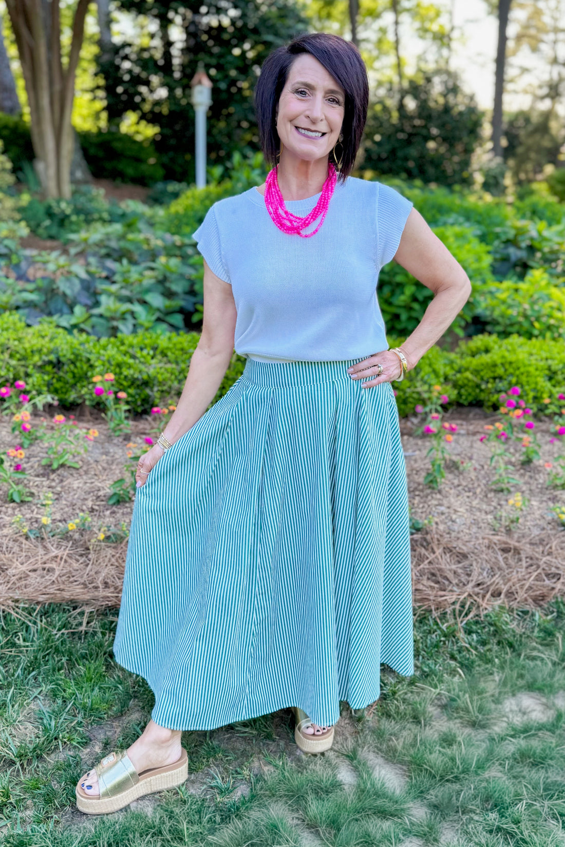 Front view of woman wearing Swagger Boutique’s Green Striped Flowy Midi Skirt with white top standing outside at a park.