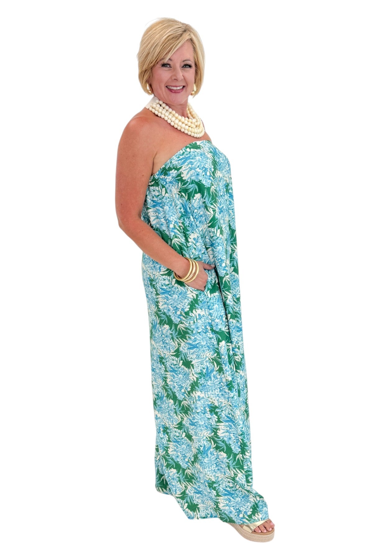 Side view of woman wearing Swagger Boutique’s Green &amp; Blue Floral Print Strapless Maxi Dress by Entro with sandals against white studio background.