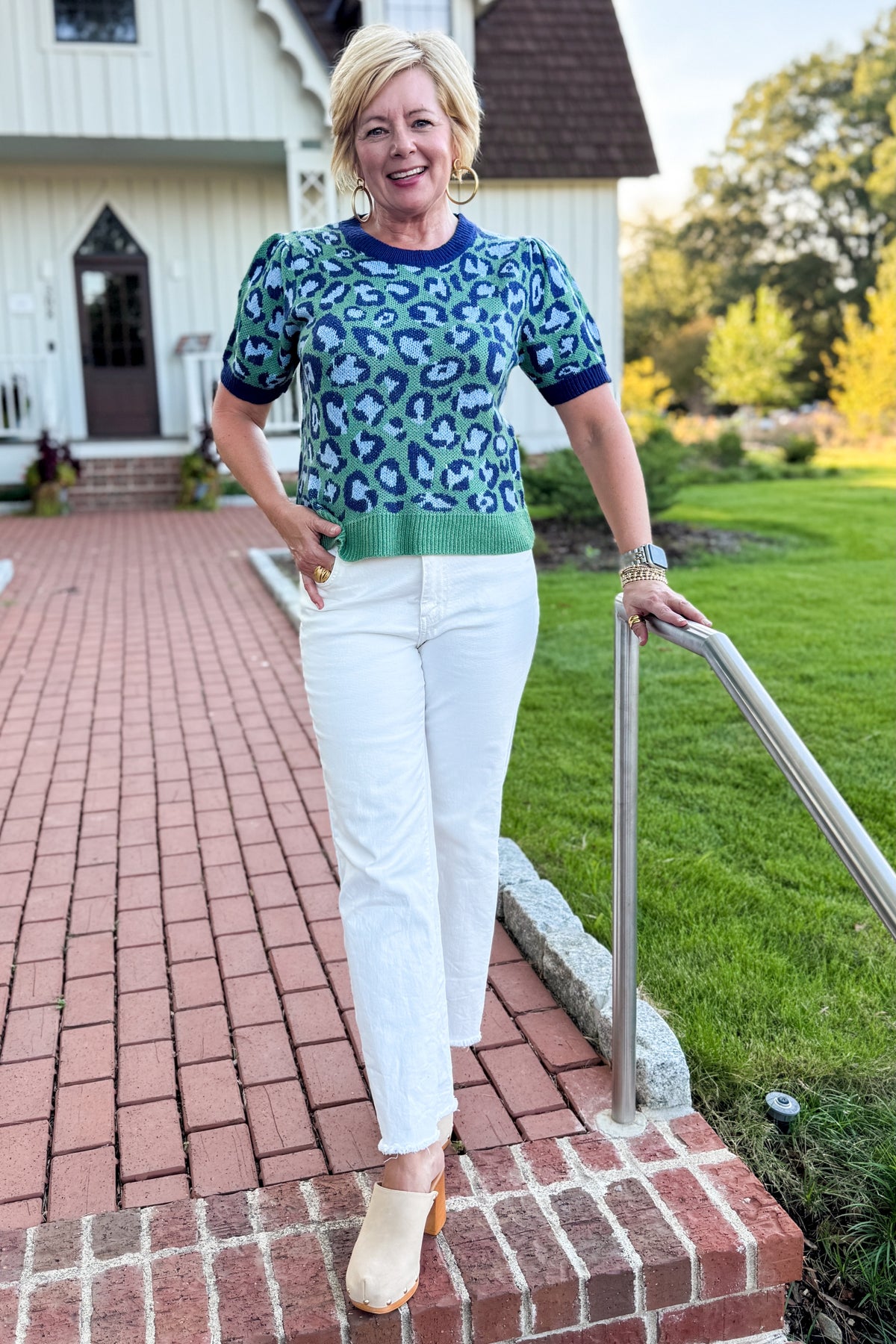 Front view of woman wearing Swagger Boutique’s Green &amp; Blue Leopard Short Sleeve Sweater with jeans standing in front of a white house.