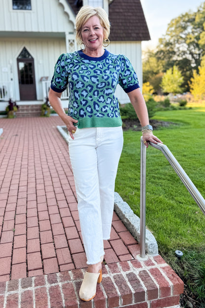 Front view of woman wearing Swagger Boutique’s Green &amp; Blue Leopard Short Sleeve Sweater with jeans standing in front of a white house.