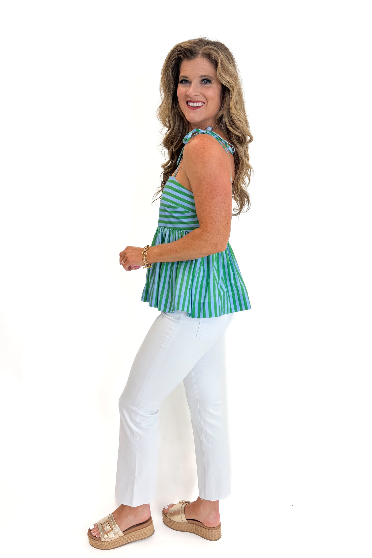Side view of woman wearing Swagger Boutique’s Green &amp; Blue Striped Peplum Tank by Entro with jeans against white studio background.