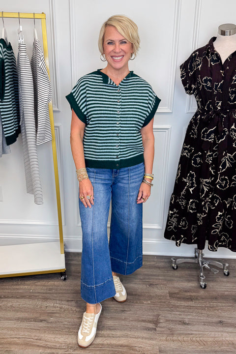 Front view of woman wearing Swagger Boutique’s Green &amp; Blue Striped Sleeveless Sweater with jeans with clothing hanging in the background.