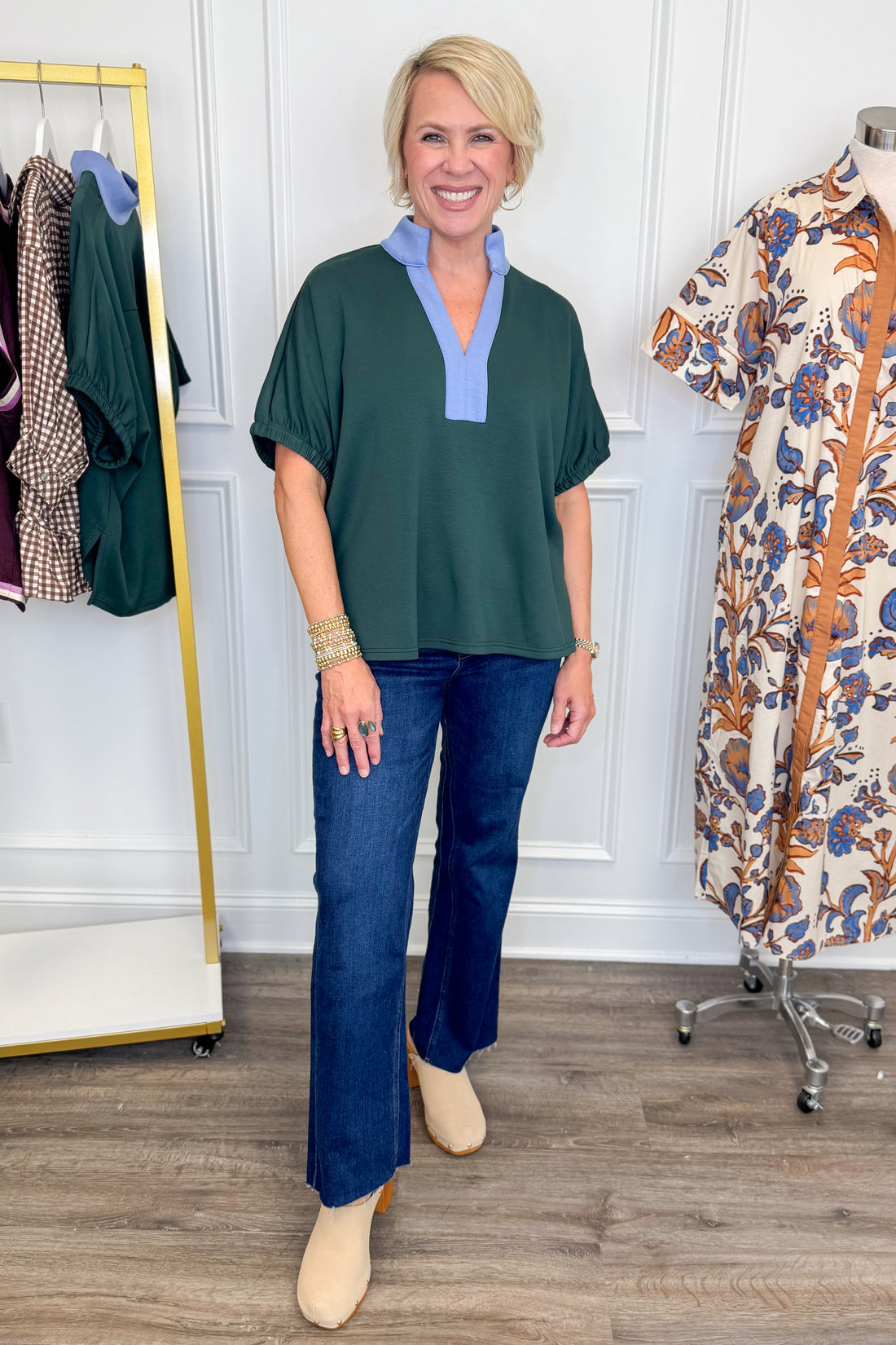A woman modeling a dark green short-sleeve blouse with a light blue V-neck collar, paired with dark blue jeans and beige ankle boots, standing in a boutique setting.