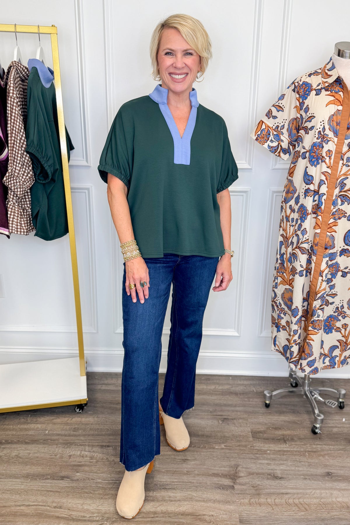 A woman modeling a dark green short-sleeve blouse with a light blue V-neck collar, paired with dark blue jeans and beige ankle boots, standing in a boutique setting.