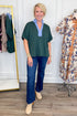 A woman modeling a dark green short-sleeve blouse with a light blue V-neck collar, paired with dark blue jeans and beige ankle boots, standing in a boutique setting.