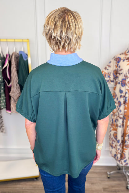 A woman viewed from behind wearing a dark teal green short-sleeve blouse with a light blue collar, paired with blue jeans in a boutique setting.