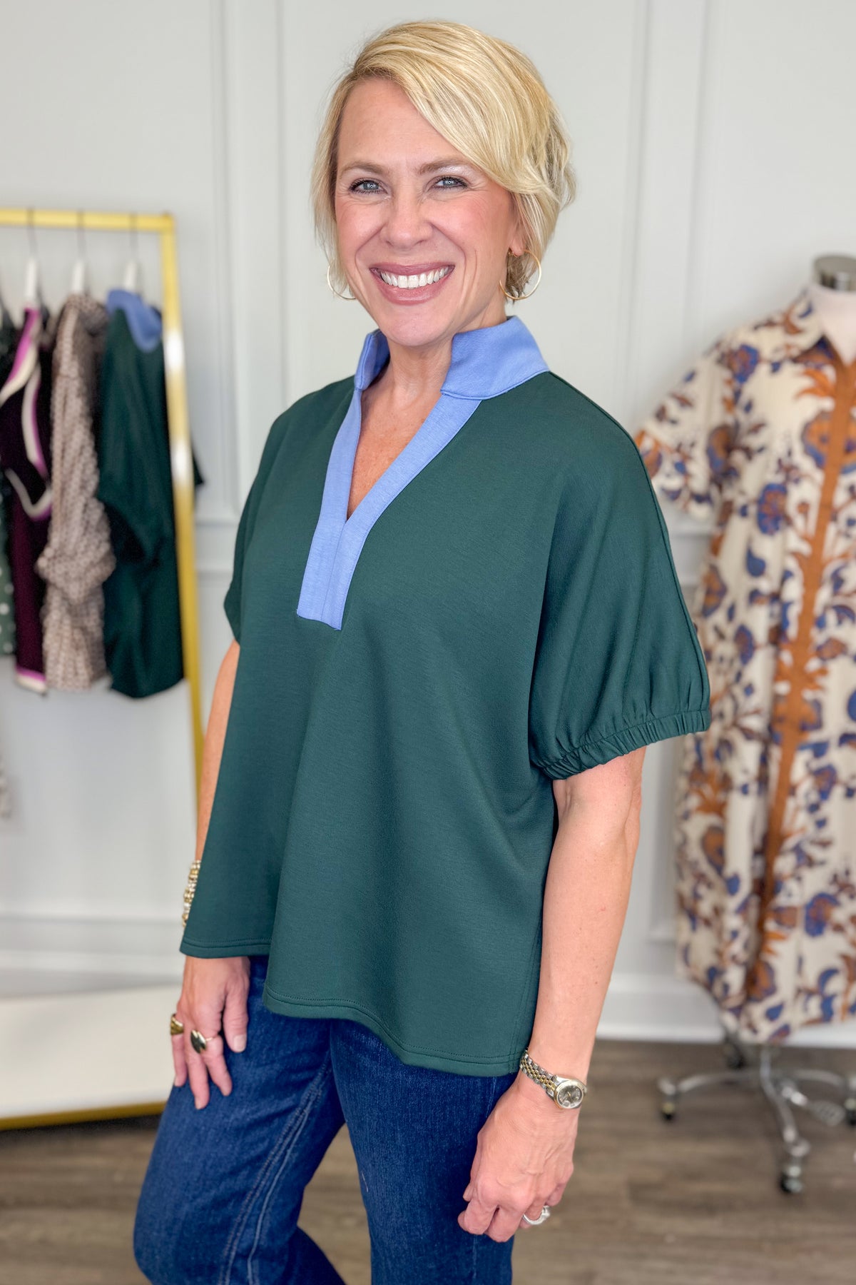 A smiling blonde woman wearing a dark green short-sleeve top with a light blue V-neck collar, paired with blue jeans, standing in a boutique setting.