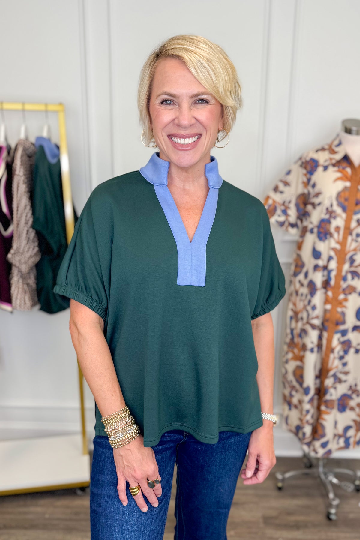 A smiling blonde woman wearing a dark green short-sleeve blouse with a light blue V-neck trim, paired with blue jeans, standing in a boutique setting.