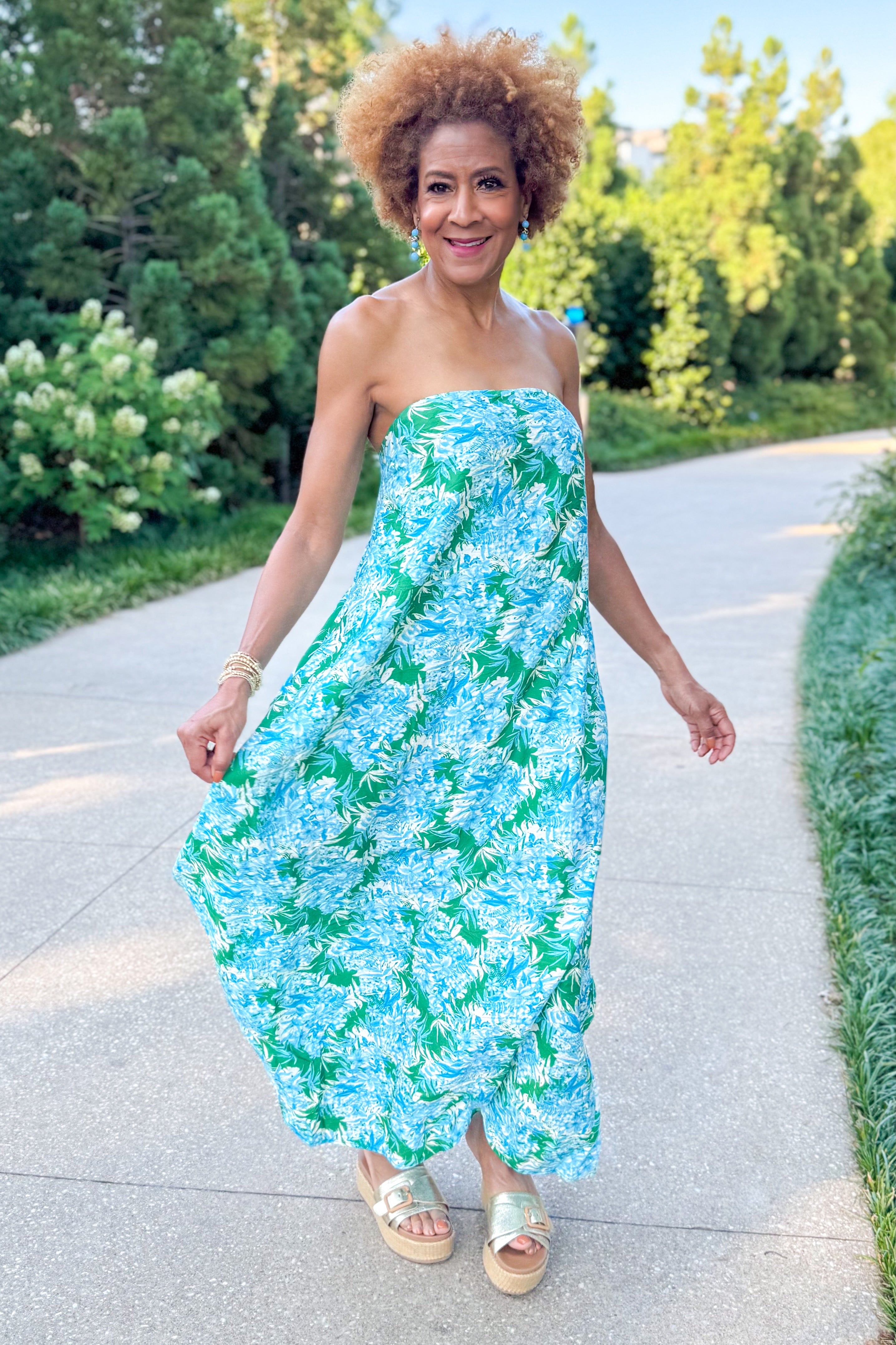 Front view of woman wearing Swagger Boutique’s Green &amp; Blue Floral Print Strapless Maxi Dress by Entro with sandals walking outside at a park.