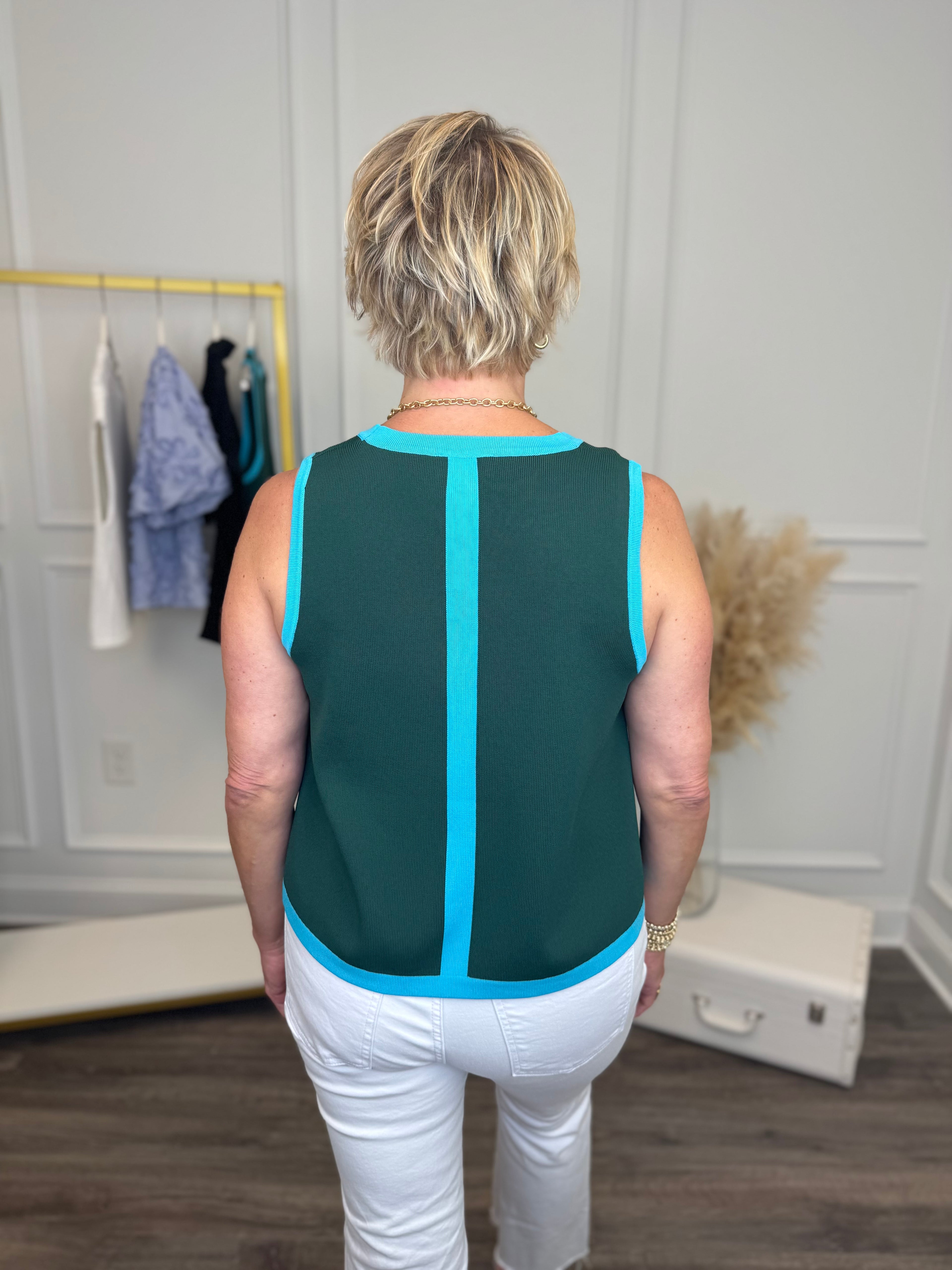 back view of woman wearing a teal sleeveless knit sweater with white jeans. 